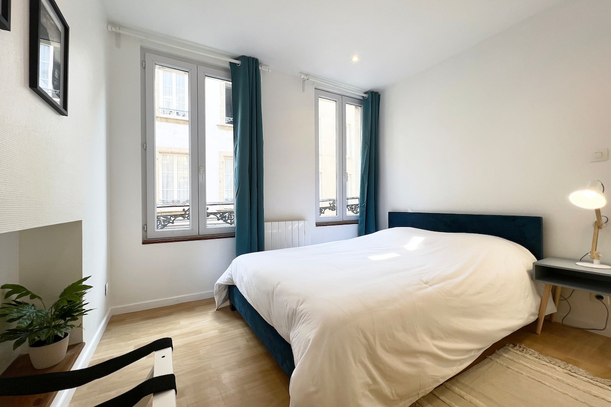 A bright bedroom features a spacious bed with a thick white comforter. Natural light fills the room through multiple windows with light blue curtains. A small lamp sits on a bedside table beside a minimalist chair, while a potted plant adds a touch of greenery.