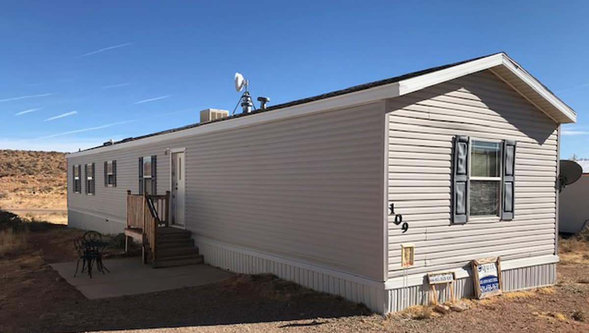 A single-story structure with light-colored siding features multiple windows framed in dark blue. A small set of steps leads up to a wooden porch. The surrounding landscape consists of dry grass and distant hills under a clear blue sky.