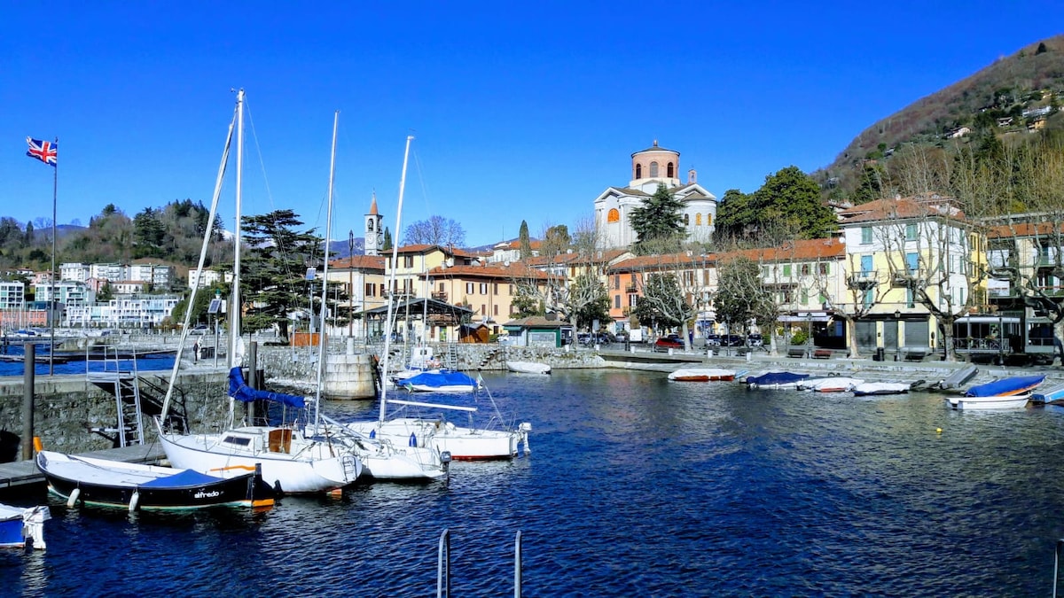 A serene lakeside view captures moored boats in calm waters, with charming buildings lining the shore. A church steeple rises among trees, while mountains serve as a backdrop, highlighting the natural beauty of the area.