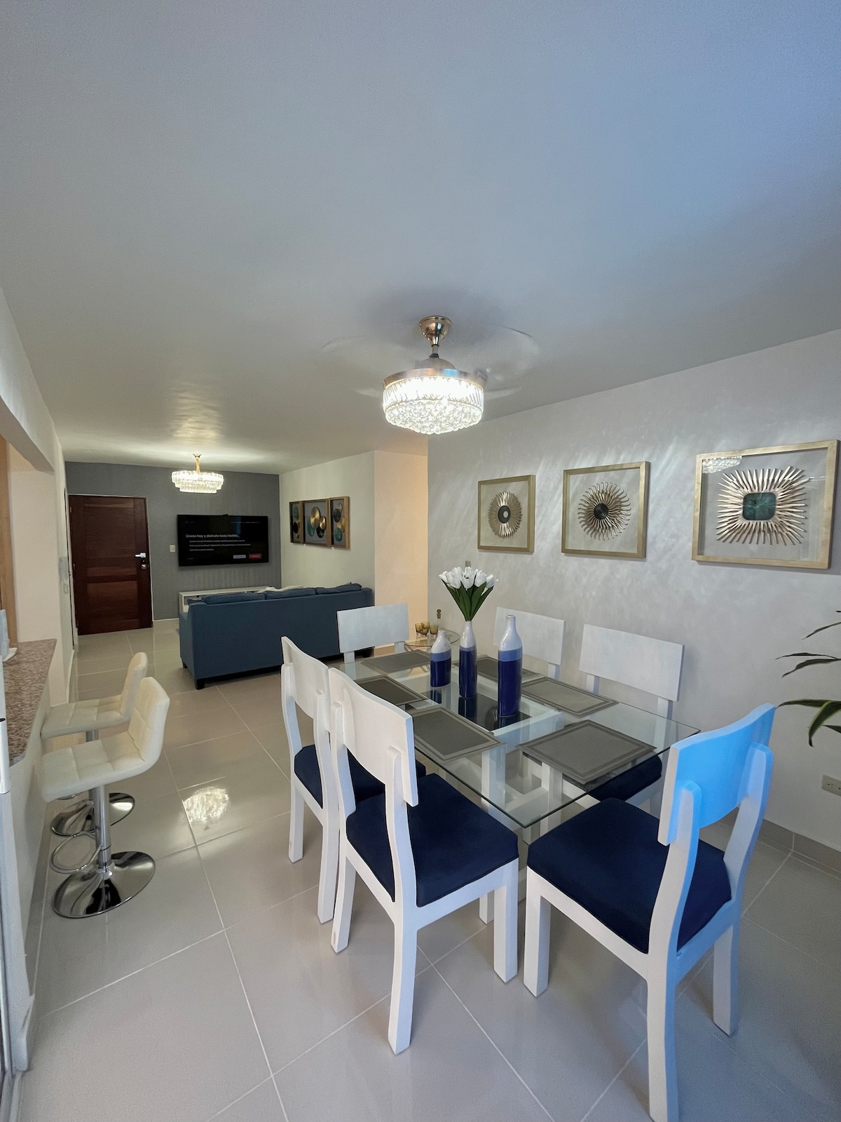 A dining area is showcased with a glass table surrounded by six white chairs, featuring blue cushions. A modern chandelier hangs above, illuminating the space. A seating area with a blue sofa is visible in the background, along with a wall-mounted flat-screen TV.