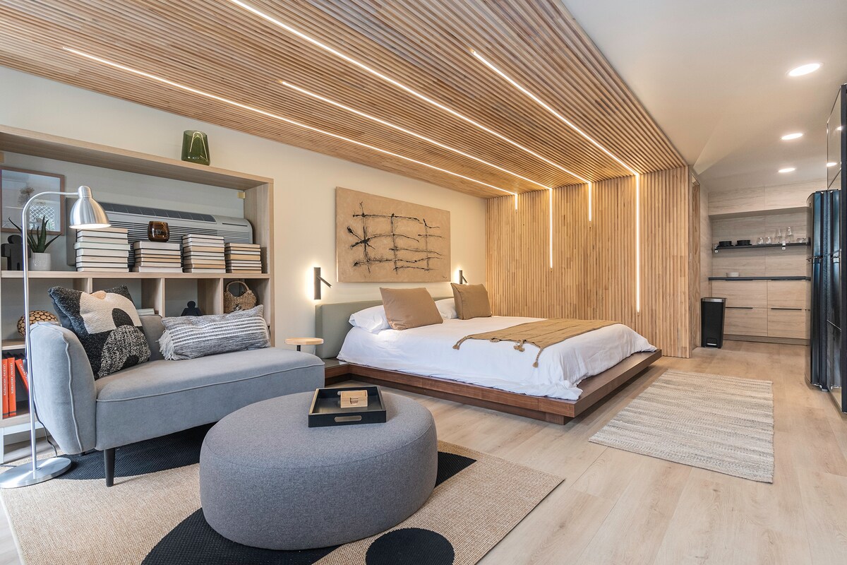 A modern studio features a spacious bed with neutral bedding, complemented by warm wood paneling and unique ceiling lighting. A cozy seating area includes a soft sofa and round ottoman, creating an inviting space. The kitchenette with sleek cabinetry is located in the background.