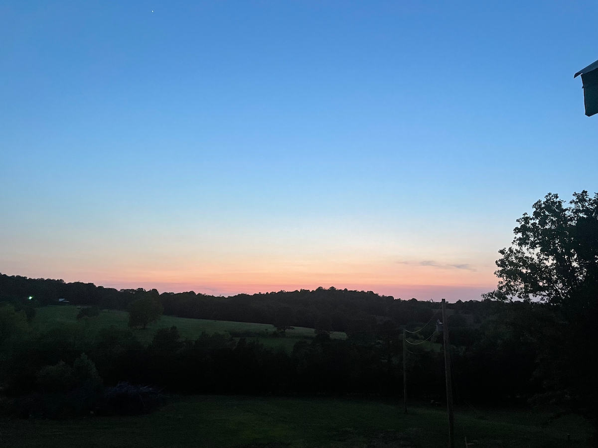 The image captures a serene landscape at dusk, showcasing a gradient sky transitioning from soft blue to gentle peach hues. Silhouetted trees and rolling hills create a tranquil foreground, while distant lights hint at neighboring properties. The peaceful ambiance is enhanced by the unobstructed views.