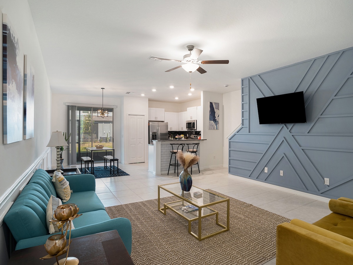 The living area features a modern layout with a turquoise sofa, stylish coffee table, and an abstract accent wall. A ceiling fan provides airflow, while a mounted television adds entertainment options. The open space leads to the kitchen and dining area, creating an inviting atmosphere.