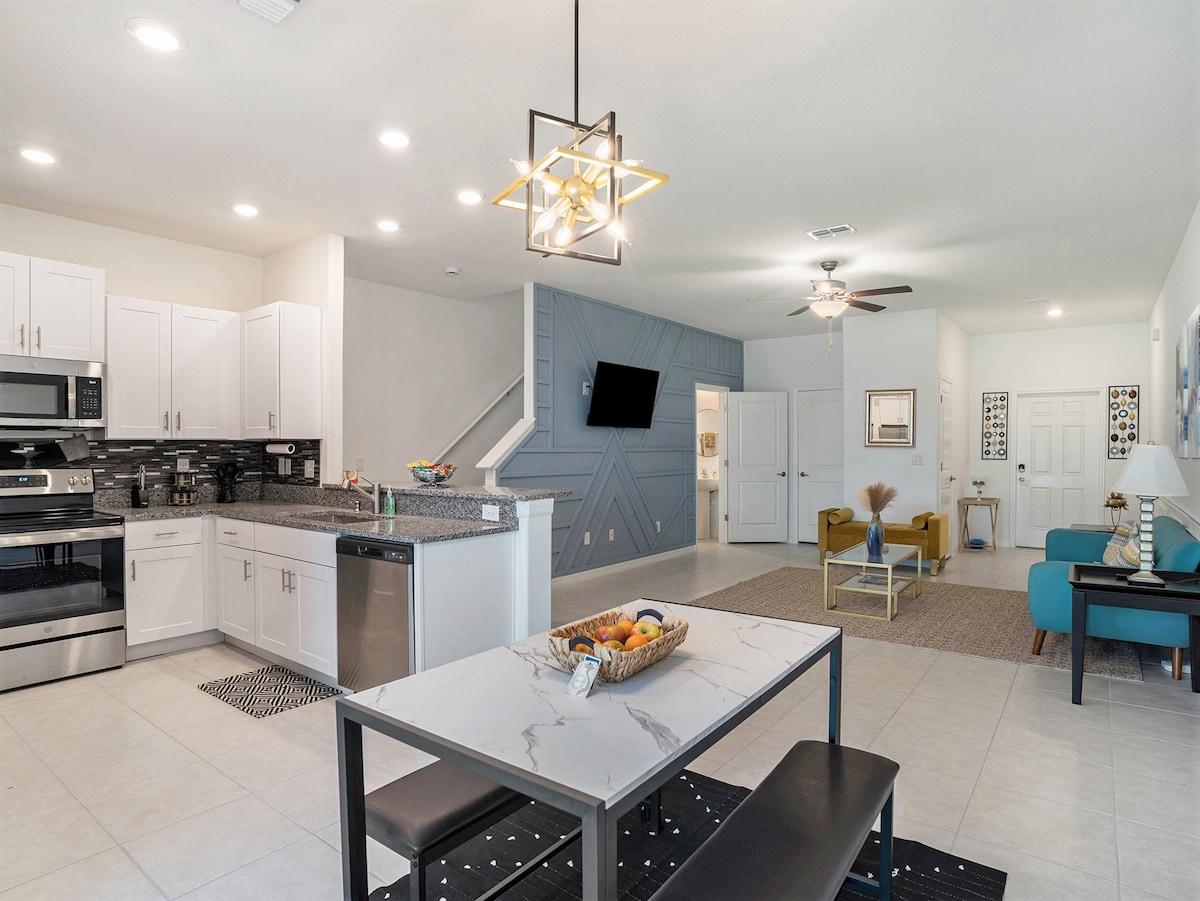 A modern kitchen and dining area showcases stainless steel appliances and a marble dining table with bench seating. Natural light illuminates the space, highlighting the sleek cabinetry and abstract accent wall. Comfortable seating is positioned nearby, creating a welcoming open floor plan.
