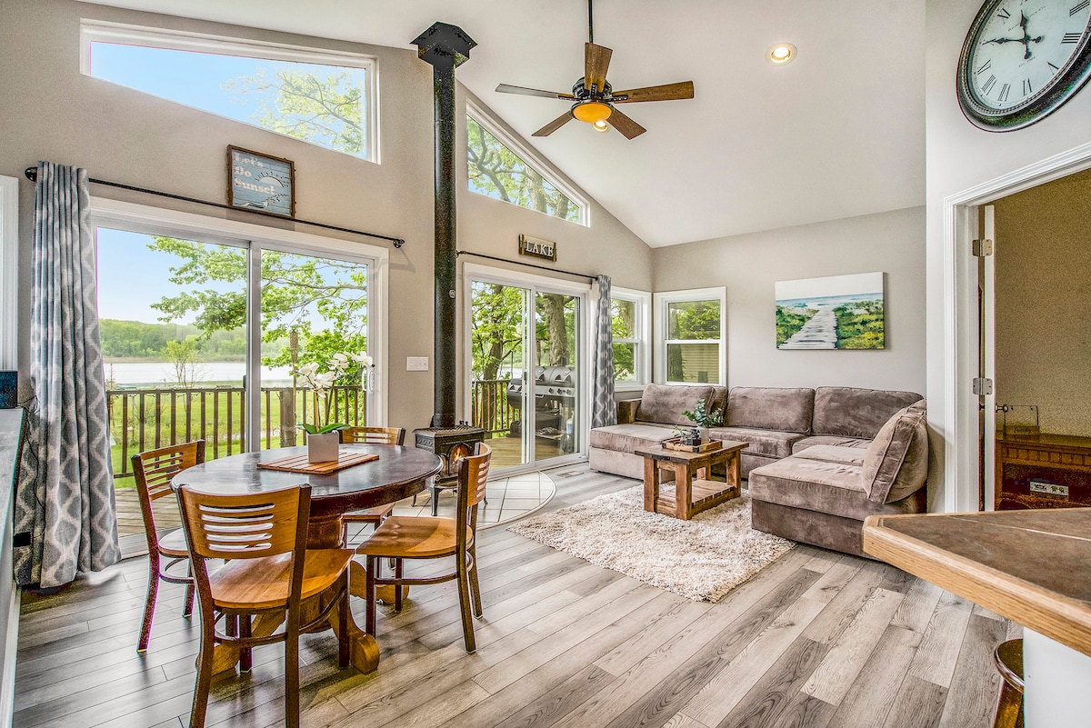 A spacious living area is highlighted by large windows that provide ample natural light and views of the surrounding landscape. A comfortable sectional sofa and a wooden dining table with seating for four are positioned on a hardwood floor, enhancing the welcoming environment.