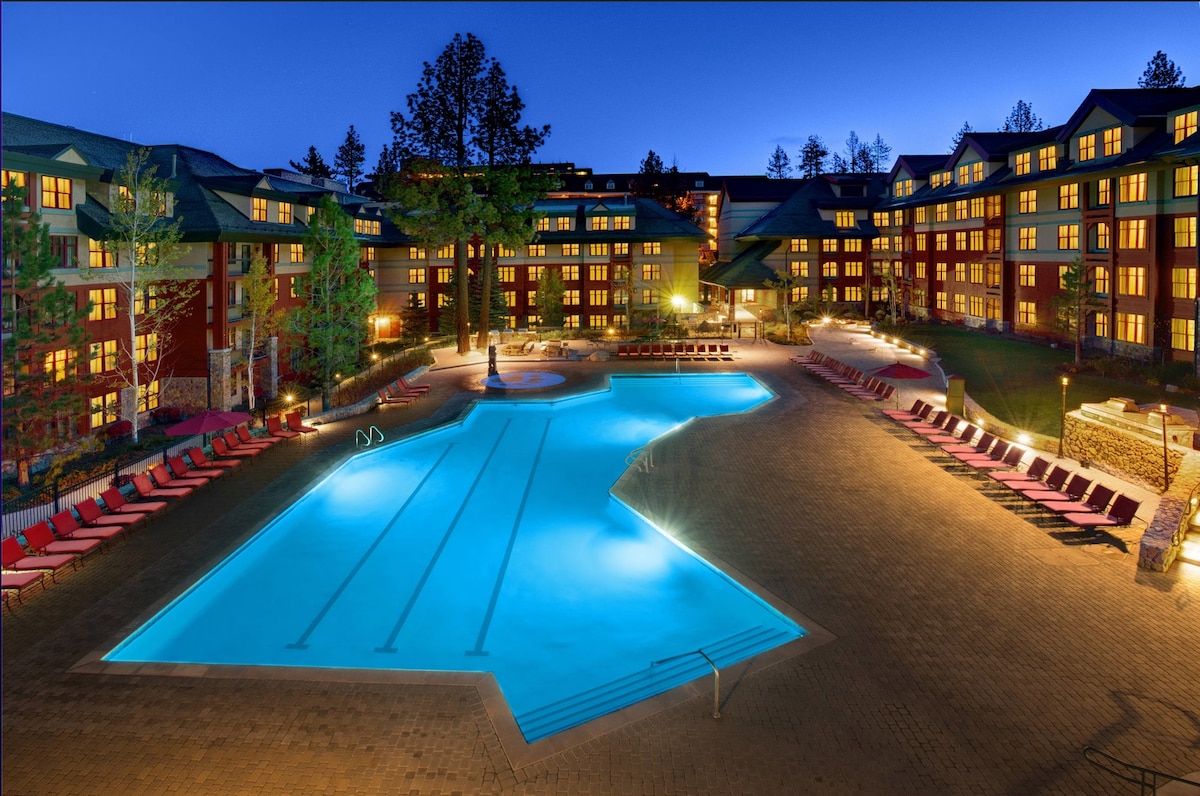The outdoor heated pool is illuminated at night, surrounded by lounge chairs. The well-landscaped resort is visible in the background, featuring multiple floors with numerous windows, reflecting warm light. The serene atmosphere is complemented by tall trees nearby.