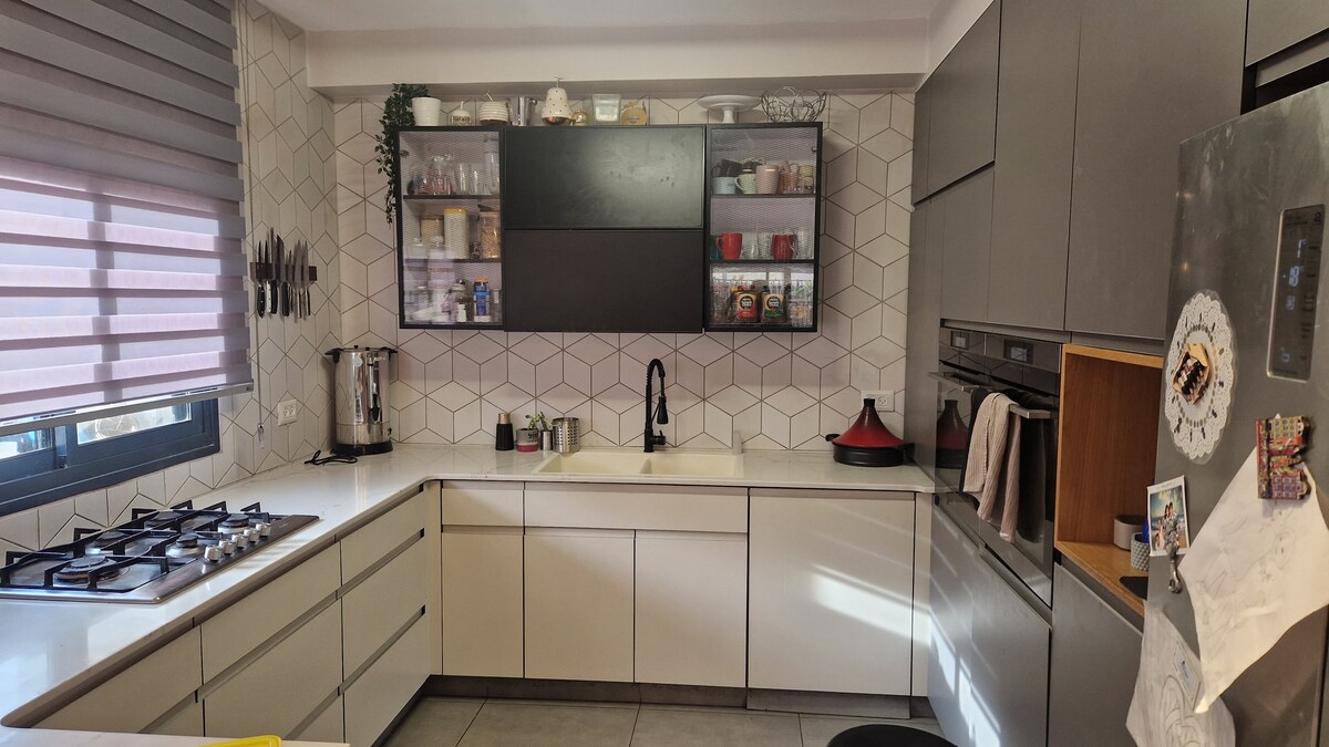 The kitchen features modern cabinetry with a sleek design, complemented by geometric tile backsplash. A spacious counter hosts a stove and sink, while glass-fronted cabinets display various kitchen items. Natural light filters in through a window, enhancing the functional layout.