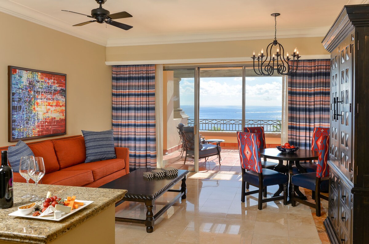 A well-appointed living area features an orange sofa paired with blue accent pillows. A dark wood table and chairs are positioned nearby, with a scenic view of the Pacific visible through large glass doors. Natural light floods the space, highlighting the elegant decor.