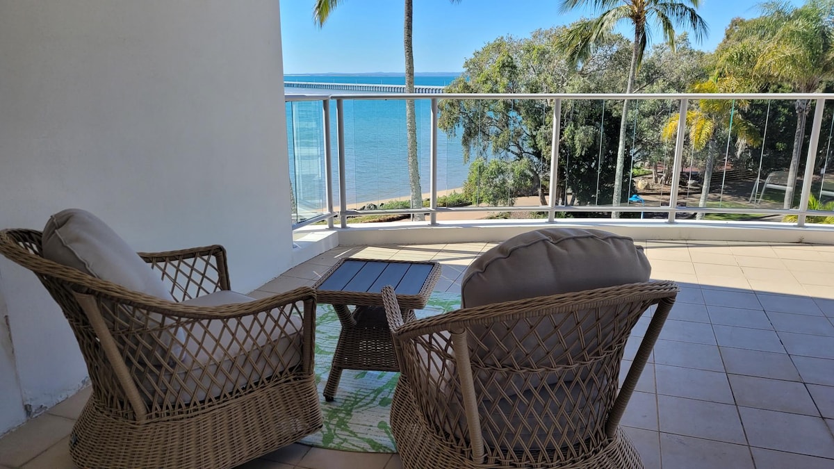 A spacious balcony features two comfortable wicker chairs positioned around a small table. The ocean is visible in the background, framed by lush palm trees. The serene setting encourages relaxation, with sunlight illuminating the area.
