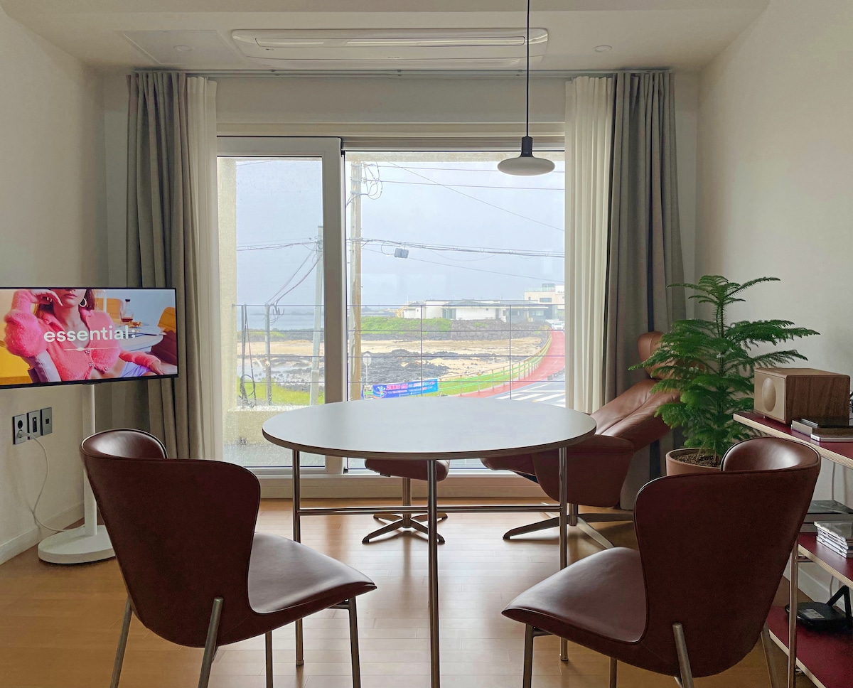 A dining area is displayed with a round table surrounded by four chairs. A wall-mounted TV is visible, showcasing content. Large windows allow natural light in, offering a view of the outside landscape, while plants add a touch of greenery to the space.