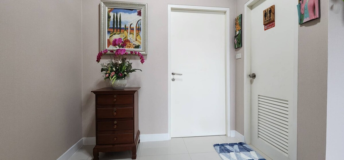 A welcoming entrance area features a wooden chest of drawers alongside a fresh floral arrangement. Colorful artwork is displayed on the walls, and a door leads to another room. A checkered mat lies on the tiled floor, offering a hint of warmth to the space.
