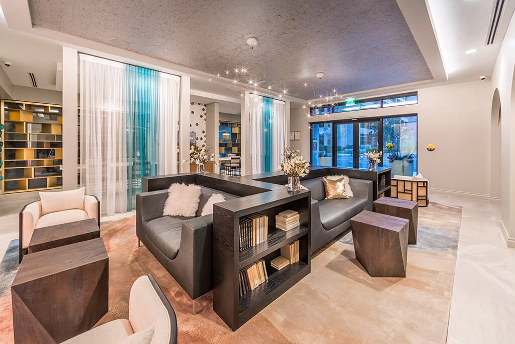 A modern social lounge is highlighted by a mix of dark wood and light upholstery furniture. The space includes low-rise seating arranged around coffee tables, with decorative books and vases placed on shelves and tables. Soft lighting and sheer curtains create an inviting atmosphere.