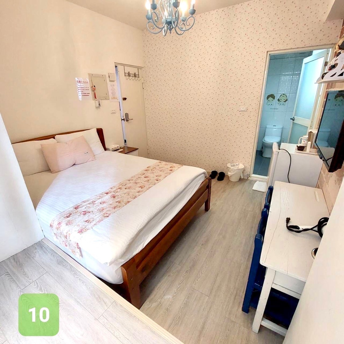 A bright room features a spacious bed with a floral accent, positioned against a patterned wall. A small desk and chair are situated on one side, with a television mounted on the wall. An en-suite bathroom is visible through a door at the rear.