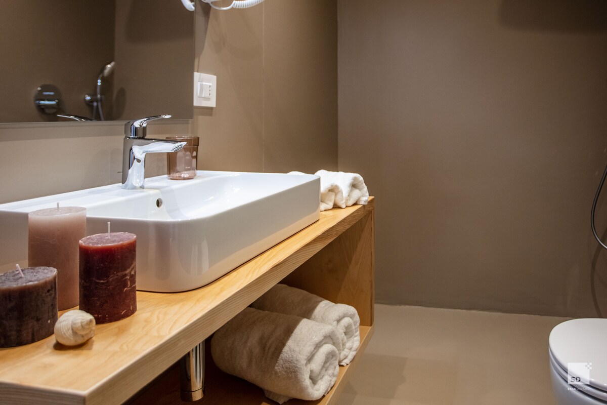 A modern bathroom features a sleek, white sink set on a wooden countertop, accompanied by three decorative candles in varying shades of brown. Soft, folded towels are neatly placed on a lower shelf, and the space is illuminated by subtle lighting.