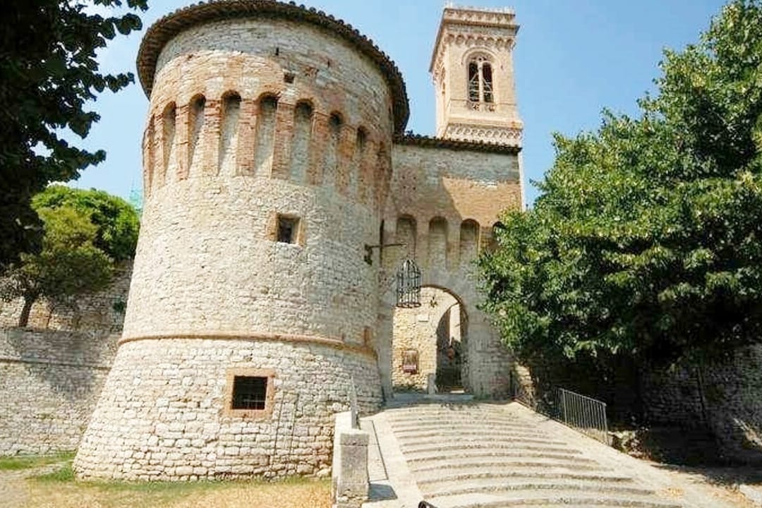 A historic stone structure features a rounded tower with a bell tower visible above. The entrance is approached via a gently sloping staircase, flanked by trees. The soft tones of the brick and stone create a blend with the natural surroundings, reflecting the rich history of the area.