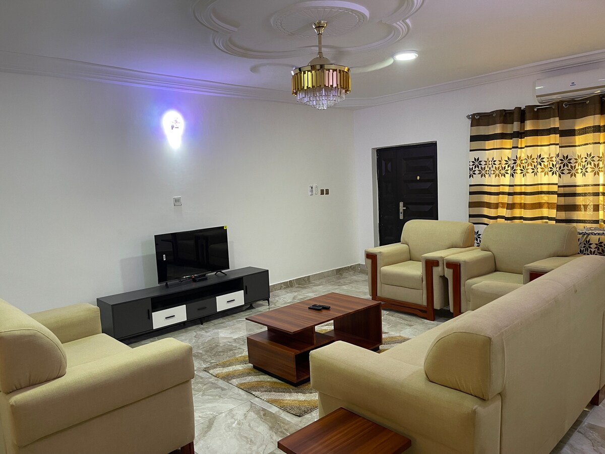 A spacious living area is highlighted by a contemporary chandelier, soft beige sofas, and a contrasting dark coffee table. A flat-screen television is situated against a wall, with light streaming in through large, patterned curtains that frame the window.