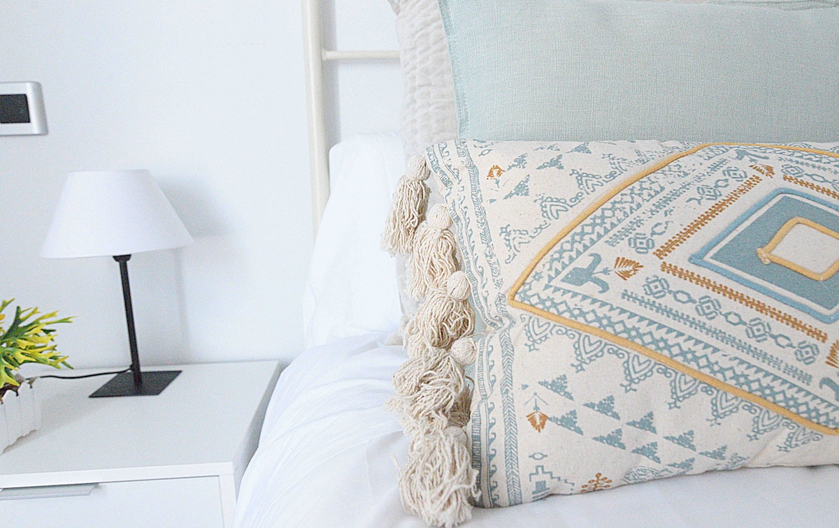 A neatly made bed features soft, layered pillows in soothing colors. A textured throw pillow with tassels adds a decorative touch. A small bedside lamp stands on a simple nightstand, while a green plant in a white container introduces a hint of nature to the space.