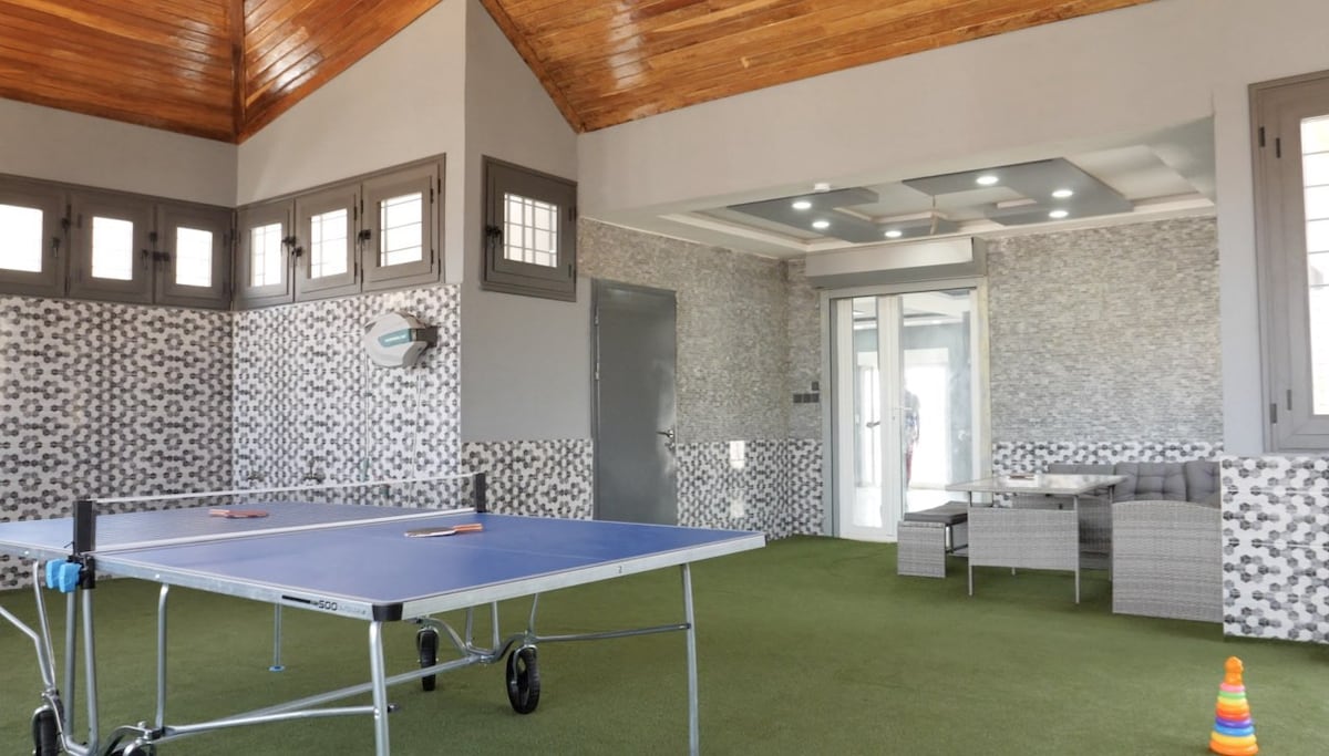 An indoor recreational area features a ping pong table set on a green carpet. Surrounding walls are adorned with patterned tiles, while large windows provide natural light. A cozy seating arrangement is positioned to the side, complemented by a wooden ceiling that enhances the vertical space.