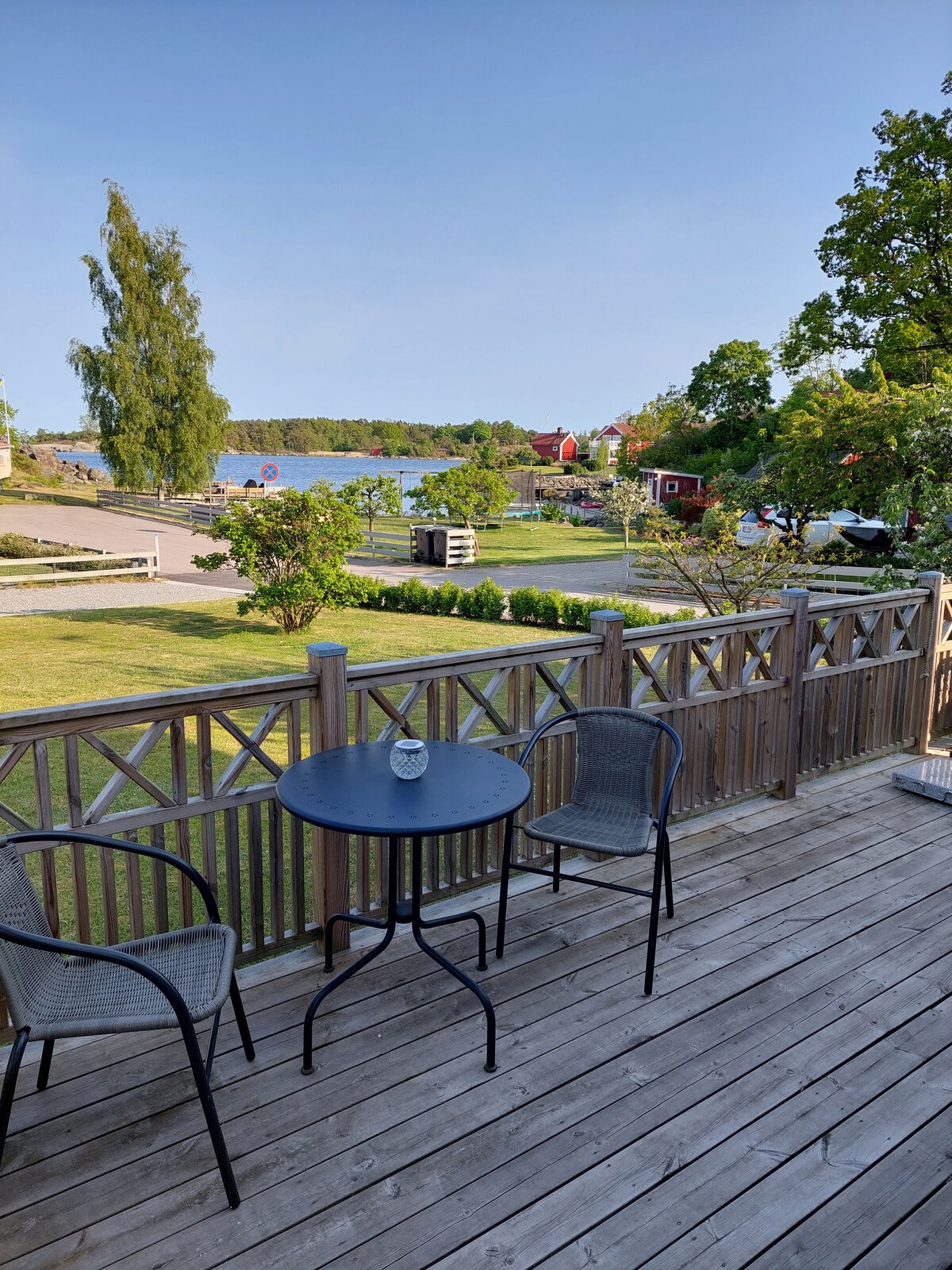 A small outdoor seating area is visible, featuring a round table and two chairs on a wooden deck. Lush greenery surrounds the space, while a view of the calm water and distant buildings can be seen beyond the railing.