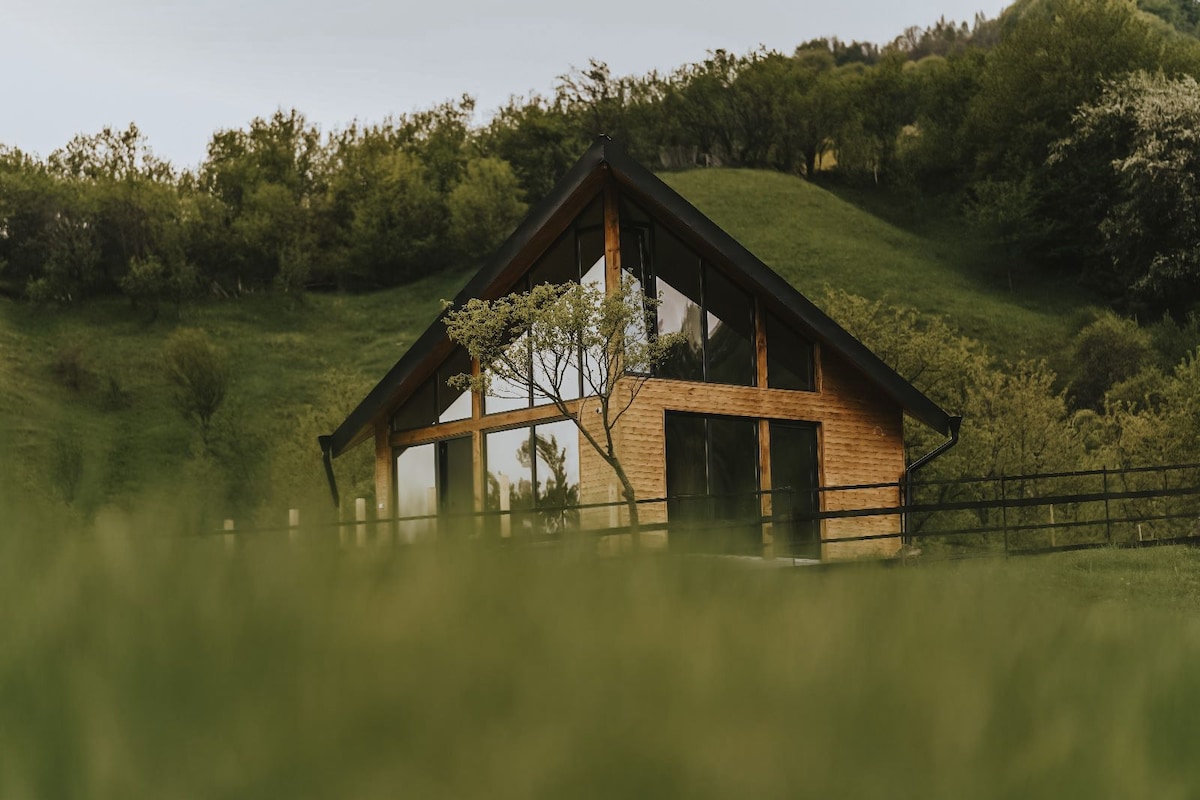 A charming cabin is set against rolling hills, featuring large glass windows that reflect the surrounding greenery. The wooden exterior complements the natural landscape, while a single tree is visible nearby, adding to the serene ambiance of the location.