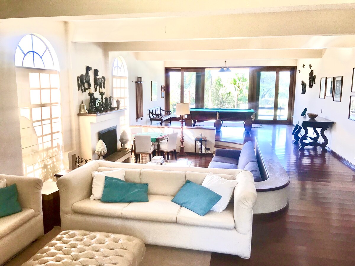 A spacious living area is highlighted by large windows providing natural light. Multiple seating areas are arranged, including a sectional couch and a tufted ottoman. A pool table is visible in the background, along with various decorative pieces adorning the walls.
