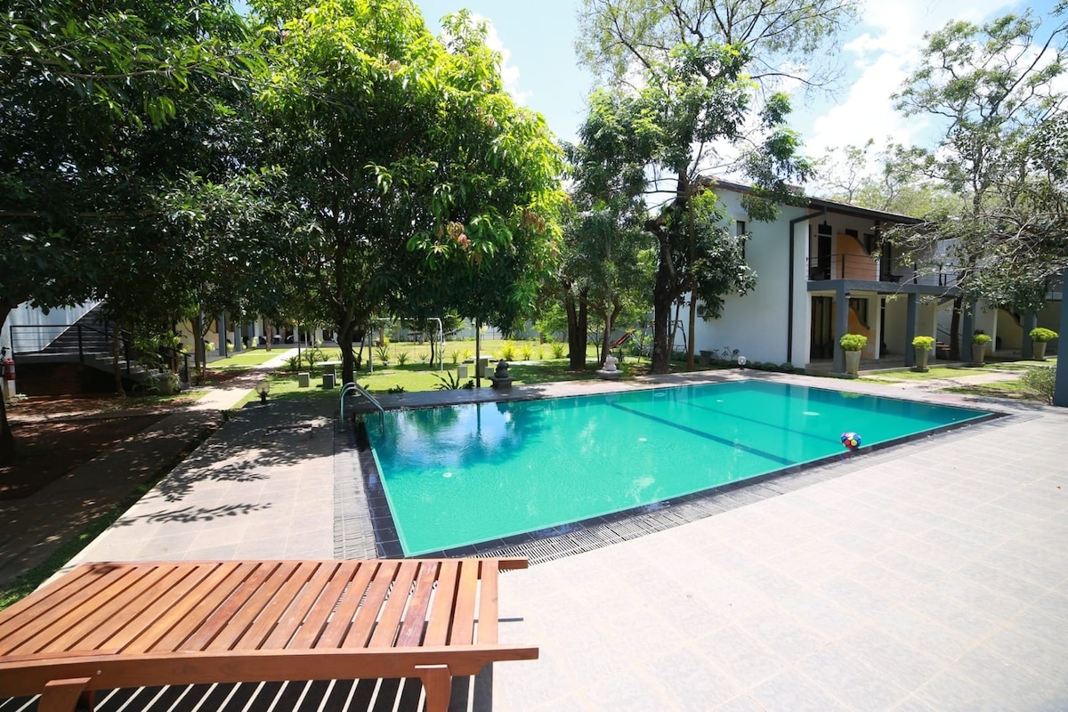 A tranquil pool area is framed by lush greenery and blooming trees, offering a refreshing escape. The clear water reflects the sky, while comfortable loungers are positioned nearby for relaxation. Surrounding structures feature a modern architectural style, contributing to a serene outdoor atmosphere.