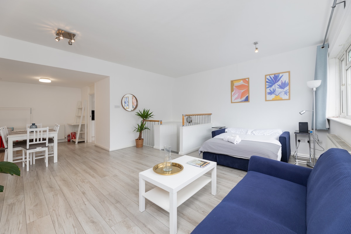 A spacious living room features light wooden flooring and an open-plan layout. A navy blue sofa, along with a white coffee table, is positioned near a double bed with a white comforter. Two framed artworks adorn the wall, complemented by an indoor plant near the staircase.