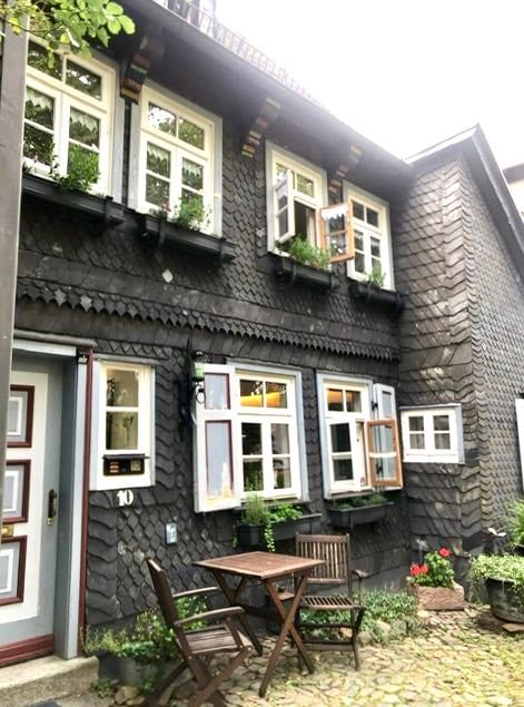 The exterior of the historic house features a unique slate facade, framed by multiple windows adorned with potted plants. A small wooden table and chairs are positioned on a stone path, inviting relaxation in the peaceful outdoor space.
