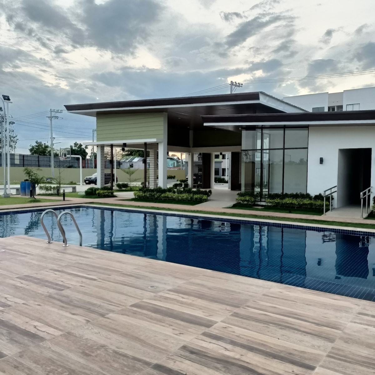 A modern swimming pool is framed by a wooden deck, with steps leading into clear water. Adjacent, a contemporary building features large glass windows and an overhanging roof. Lush greenery surrounds the pool area, enhancing the serene setting.