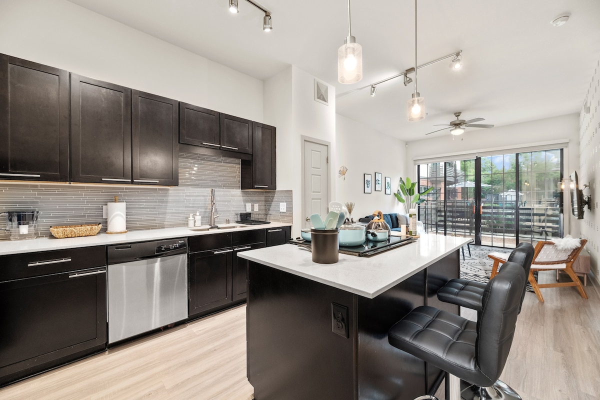 An open concept kitchen features modern black cabinetry and stainless-steel appliances. A large island with seating for two is highlighted by stylish pendant lights. Natural light floods the space through glass doors, which lead to an outdoor patio area, enhancing the inviting ambiance.