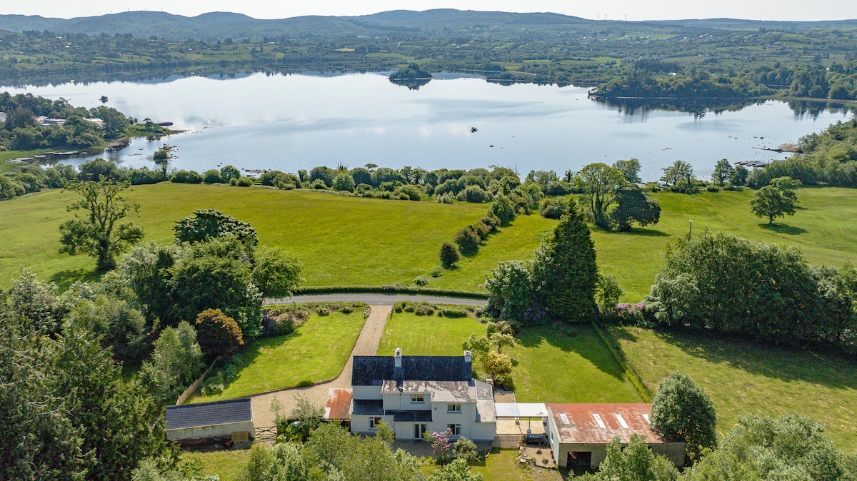 An aerial view captures the house surrounded by expansive green lawns, leading to the serene Lough Eske. The still water reflects the landscape, with trees lining the shore. Rolling hills can be seen in the distance, providing a sense of tranquility in the natural setting.