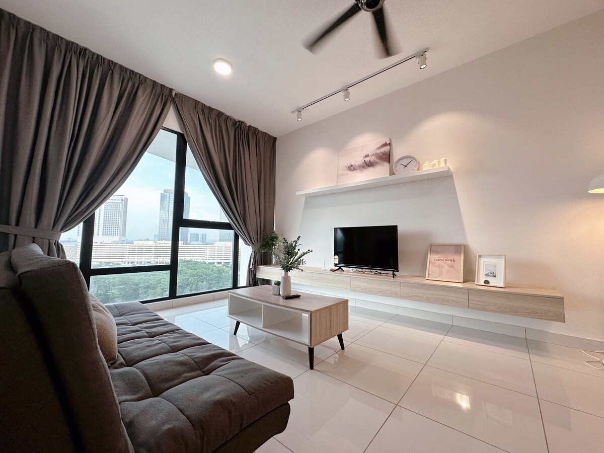 A well-lit living area features a large window with city views, allowing natural light to enter. A comfortable sofa is positioned beside a modern TV stand, where a television is set. Decorative items are displayed on the shelves, enhancing the minimalist décor.