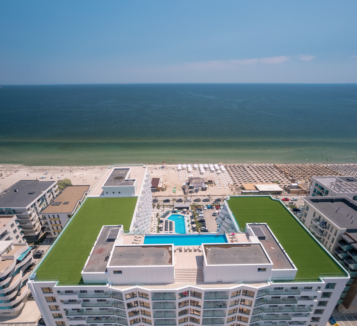 An aerial view showcases the Alezzi Infinity Aparthotel & Spa, highlighting its modern architecture and lush green rooftop. Expansive pools are visible below, bordered by sun loungers. The blue ocean and sandy beach extend in the background, providing a serene coastal setting.