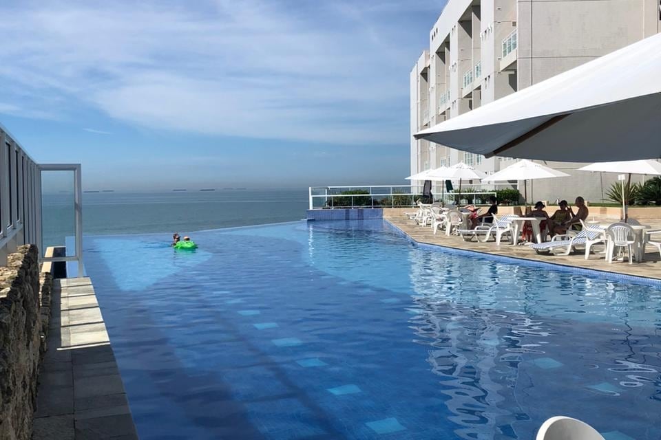 An expansive infinity pool glistens under sunlight, overlooking the open sea. Lounge chairs are arranged along the poolside, providing seating for guests. Large umbrellas offer shade, creating a comfortable space for relaxation. The serene water reflects the clear blue sky.