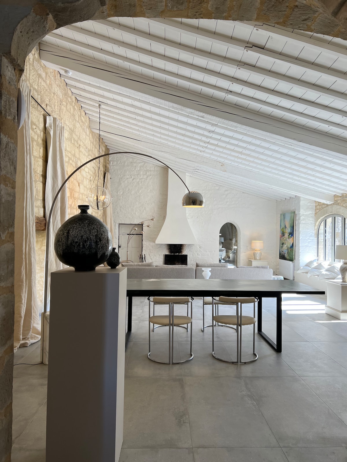 A spacious dining area features a sleek black table accompanied by two minimalist stools. A textured vase rests on a pedestal nearby. A large stone fireplace stands at the end of the room, while natural light fills the space through expansive windows.