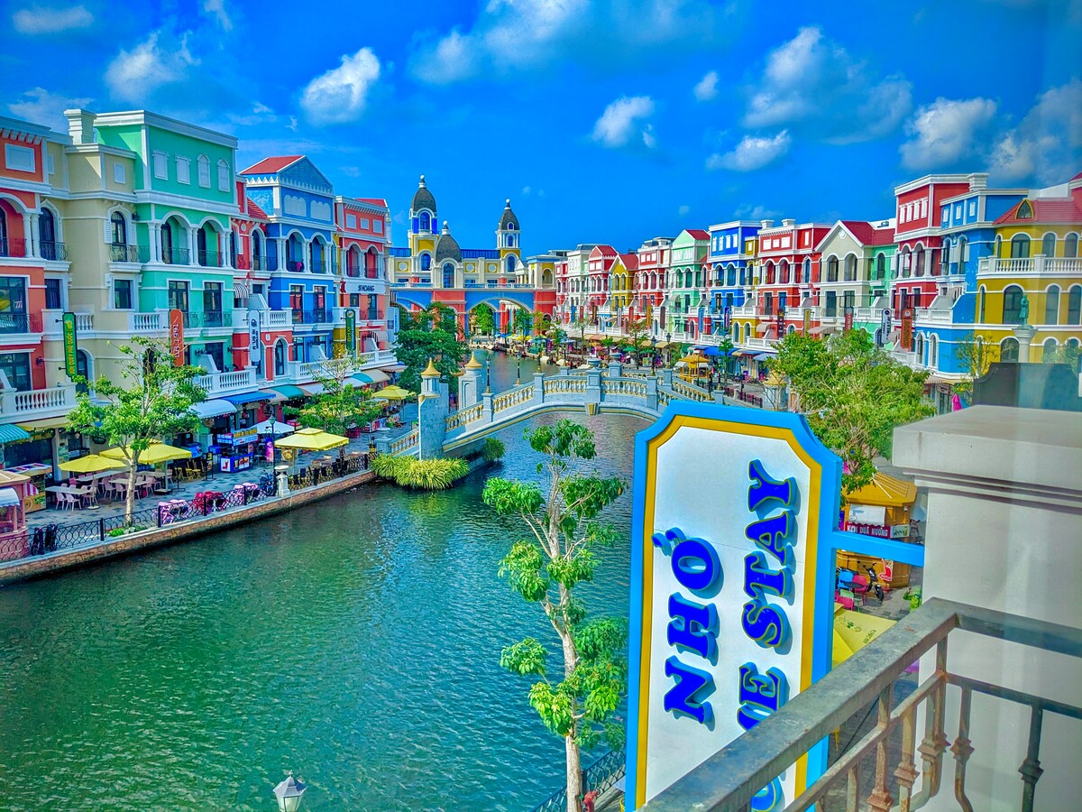 A vibrant waterfront scene is depicted, featuring colorful buildings lining a picturesque canal. Lush greenery and outdoor seating areas are visible along the water's edge, offering a lively atmosphere. Cloud-dotted blue skies complement the cheerful ambiance of the area.