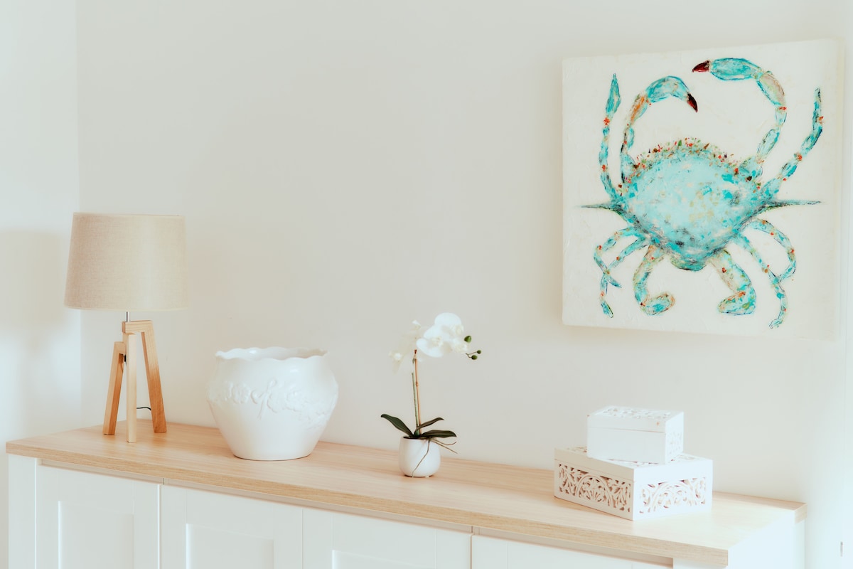 A light-colored sideboard is adorned with decorative items. A textured white vase holds a small orchid, while a larger white bowl features an intricate design. A lamp with a beige shade and a painting of a crab in turquoise tones completes the serene decor.