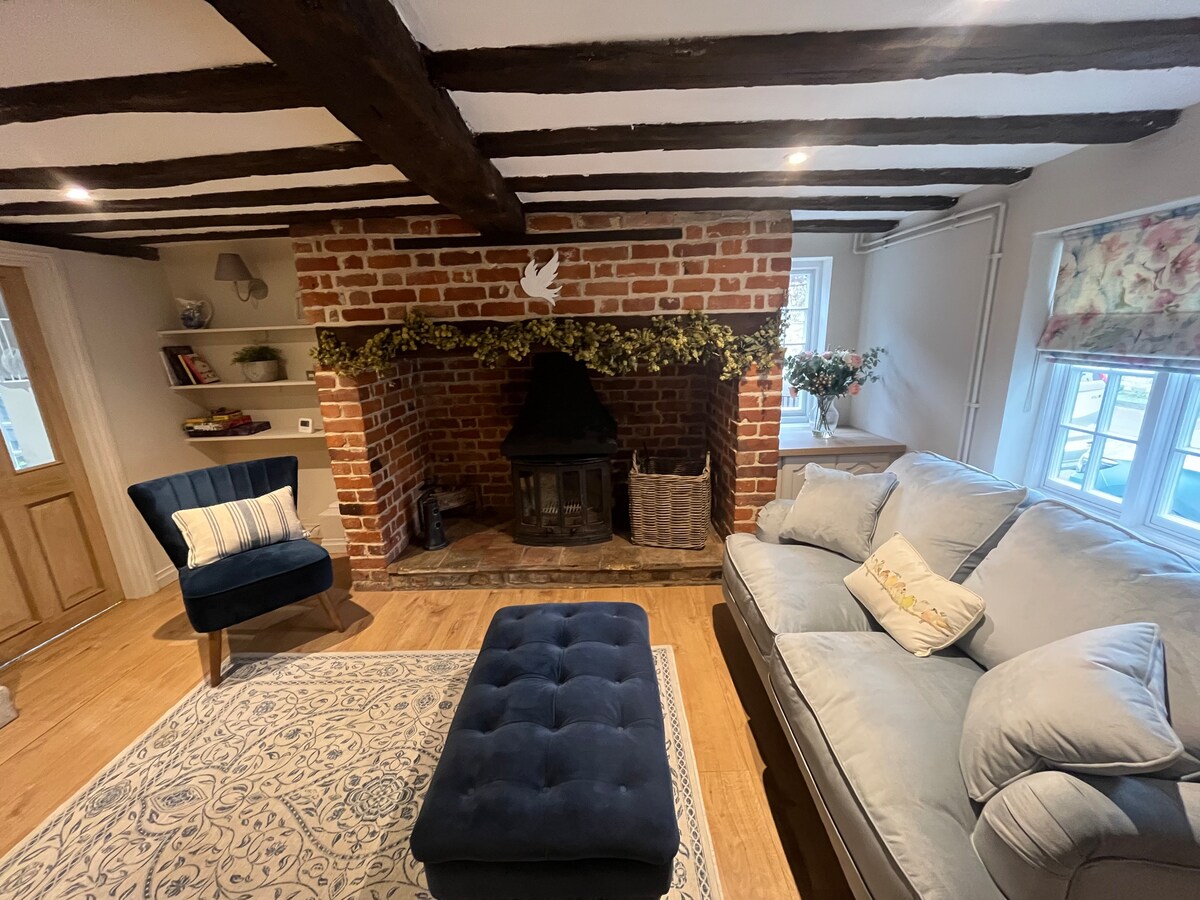 A cozy living room features a large inglenook fireplace adorned with a decorative mantle. The space includes a plush sofa and a stylish armchair, complemented by a soft area rug. Natural light enters through the window, highlighting the exposed Tudor beams above.