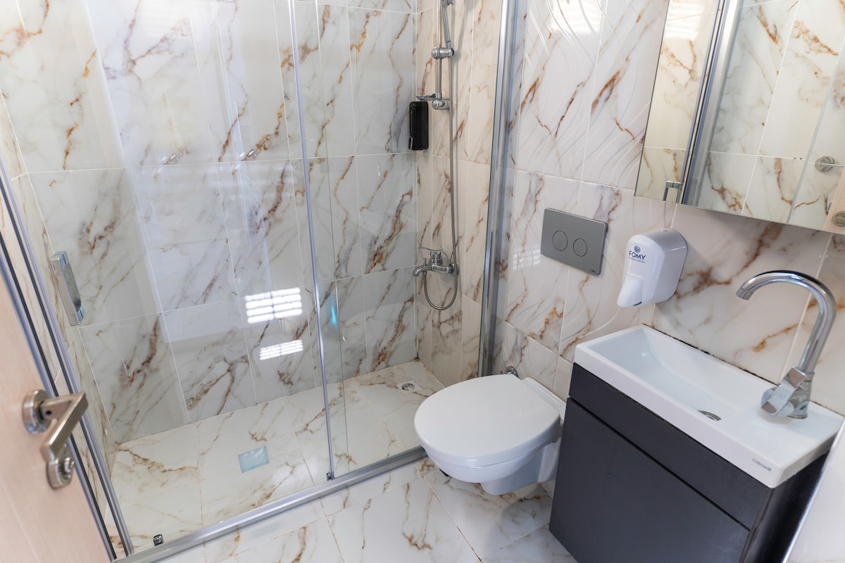 A modern bathroom features sleek marble walls and a glass-enclosed shower. A compact vanity with a sink and storage is accompanied by a toilet. Natural light filters through the space, highlighting the contemporary fixtures.