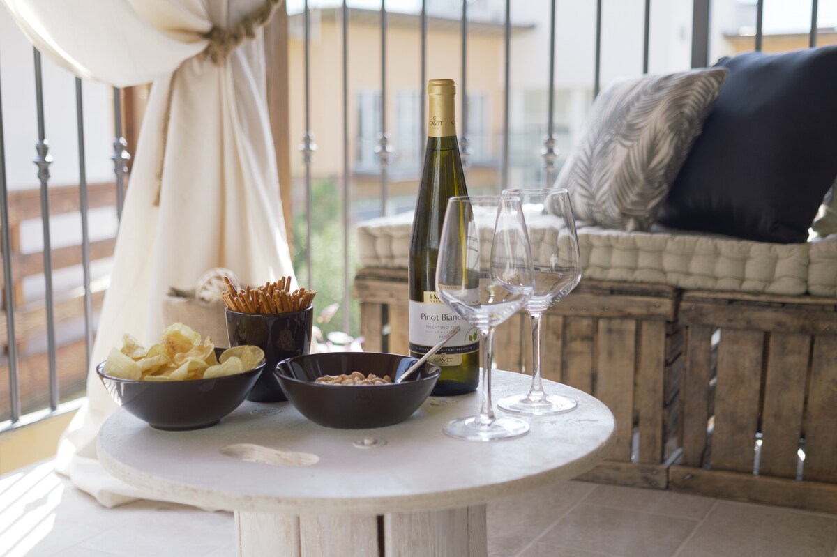 An outdoor table is set with a bottle of white wine and three wine glasses. Two bowls contain snacks: one holds potato chips, while another is filled with pretzels and nuts. A cozy cushion is visible in the background.