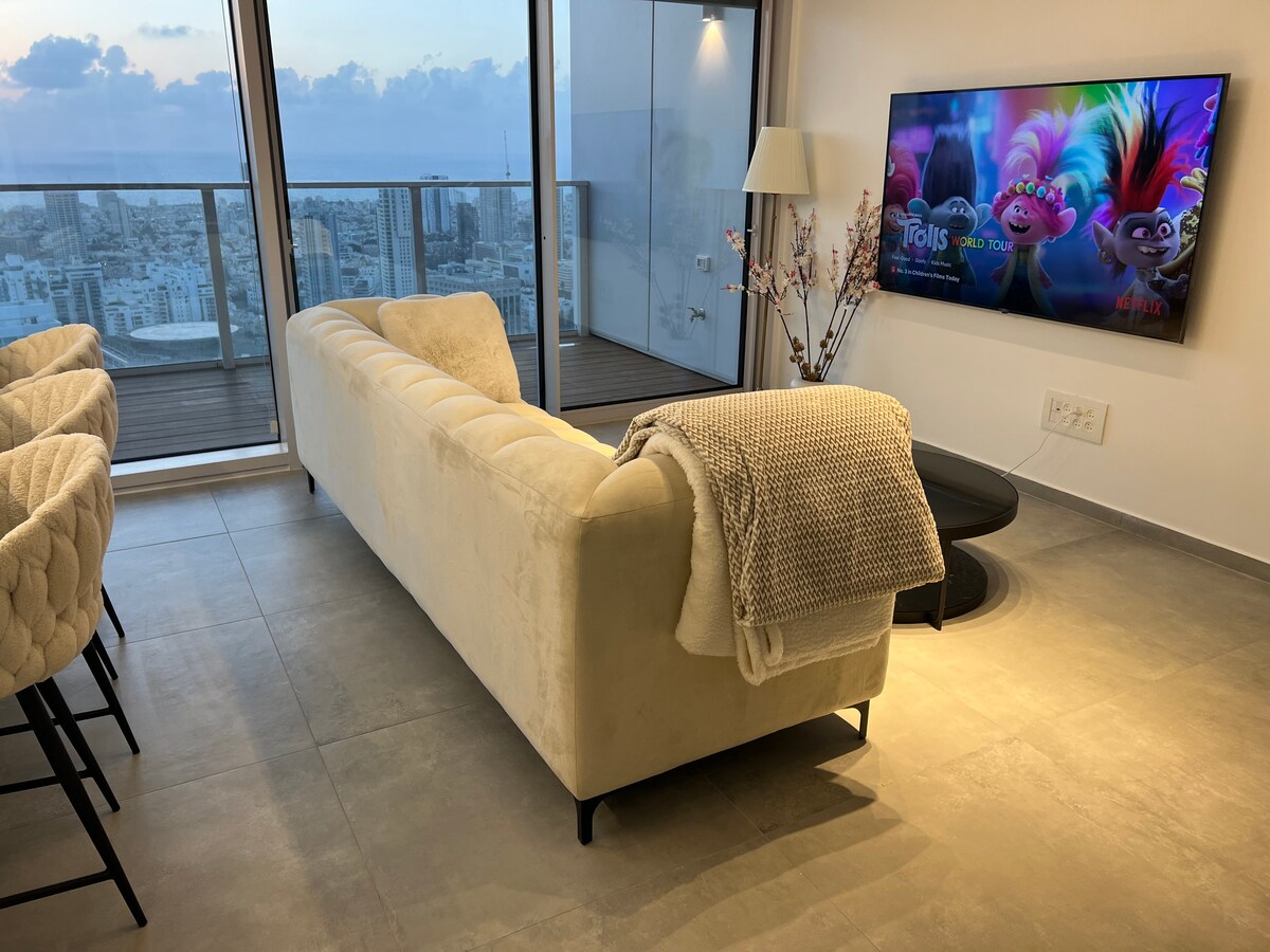 A modern living area is presented with a light-colored sofa and a cozy throw draped over the arm. A large screen displays a family-friendly film, while floor-to-ceiling windows offer panoramic views of the city skyline. Elegant bar stools are positioned at a nearby counter.