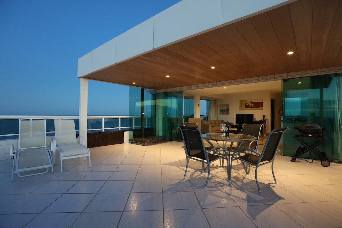 A large private terrace offers an open space with a dining table and chairs, providing views towards the ocean. Sun loungers are placed for relaxation. The area features a mix of shaded and sunlit sections, creating a versatile outdoor environment.