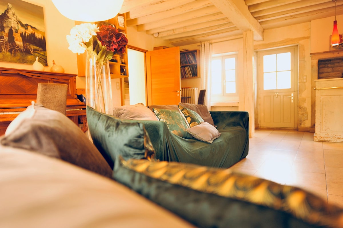 A warm living space is shown, featuring a comfortable green sofa adorned with decorative cushions. Natural light streams through a set of double doors, illuminating the room. A wooden piano and shelving units are also present, enhancing the inviting setting.
