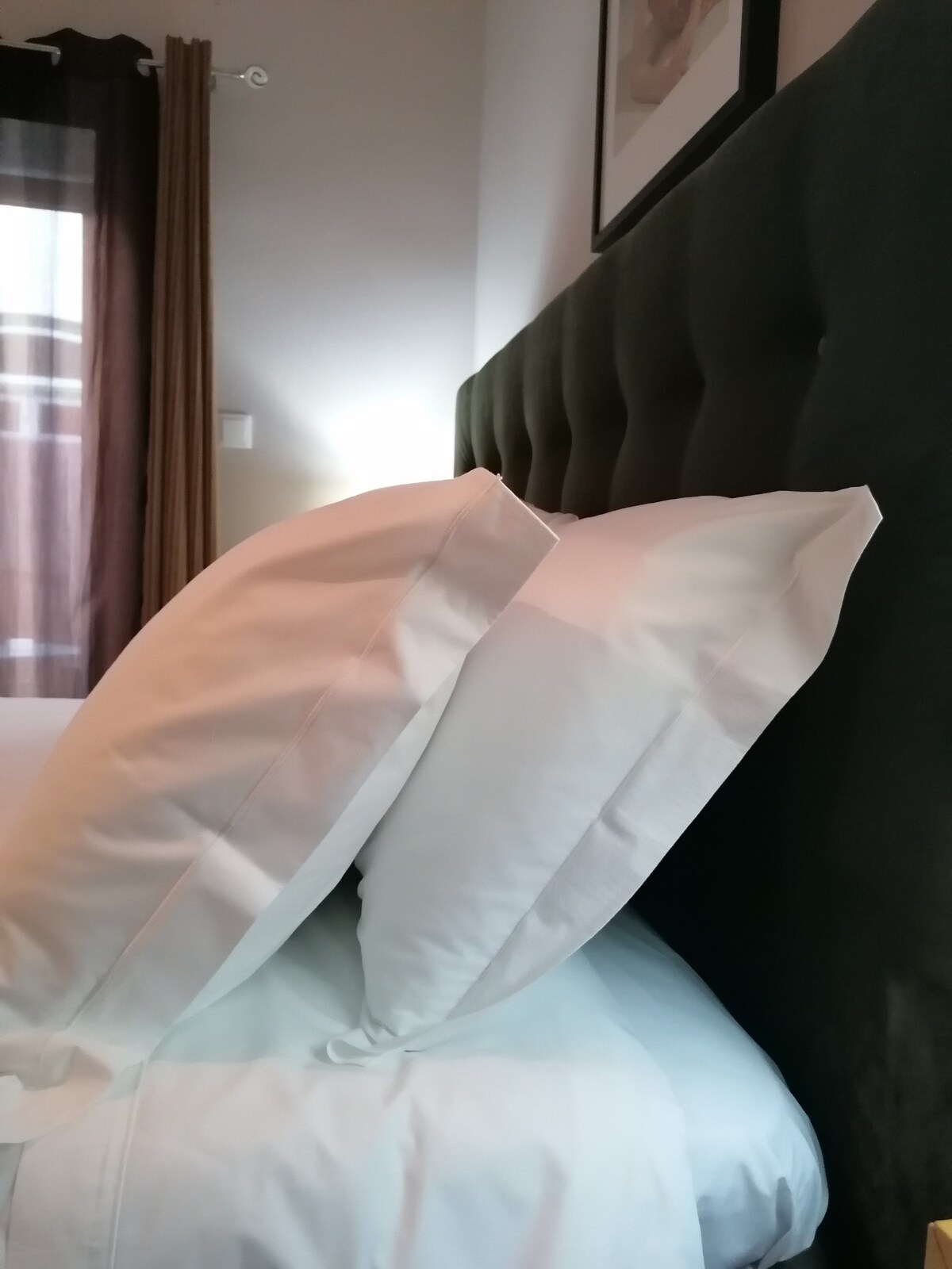 Plush pillows are neatly arranged against a tufted headboard, complementing the soft bedding. Natural light filters through sheer curtains, casting a gentle glow in the space, which is further enhanced by warm-toned wall decor.