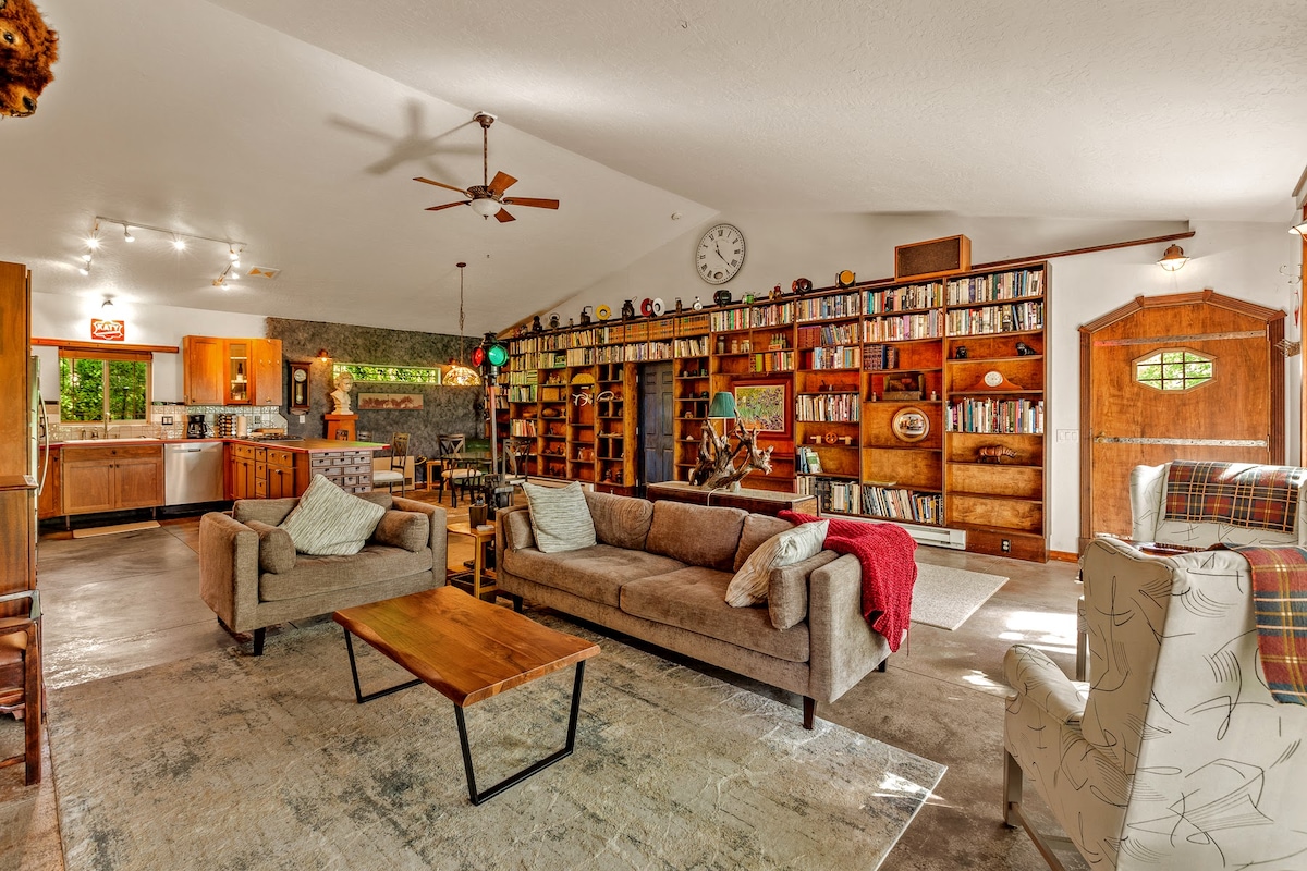 A spacious living area features a combination of soft seating arrangements and a wooden coffee table, set against a backdrop of a large bookshelf filled with various books and decorative items. A well-equipped kitchen is visible in the background, complemented by natural light streaming in through the windows.