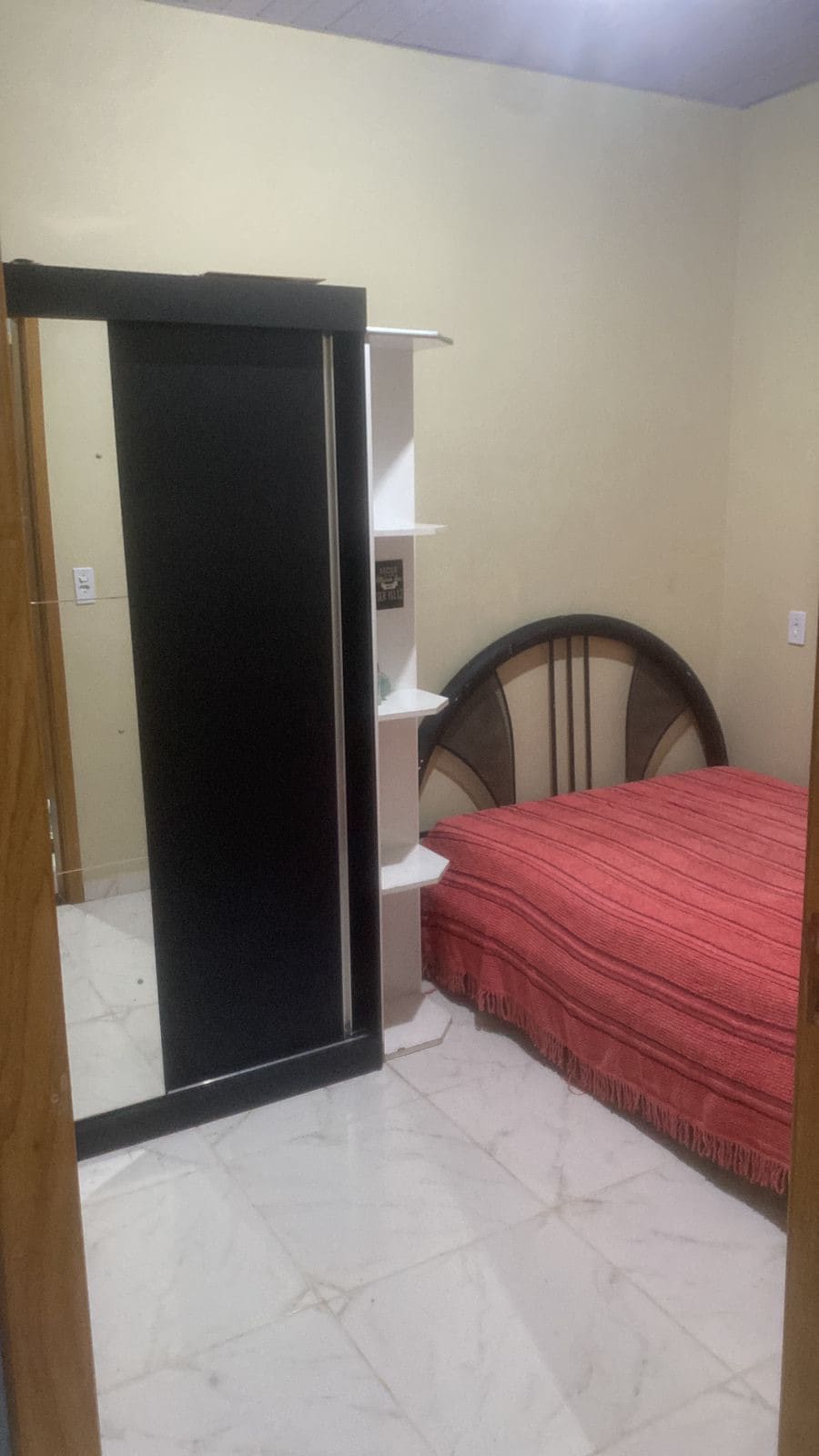 A modest bedroom features a spacious bed with a red and cream-striped blanket. A black shelving unit stands against one wall, while light-colored tiles create an open atmosphere. The walls are painted in a soft neutral tone, complementing the room's simple design.