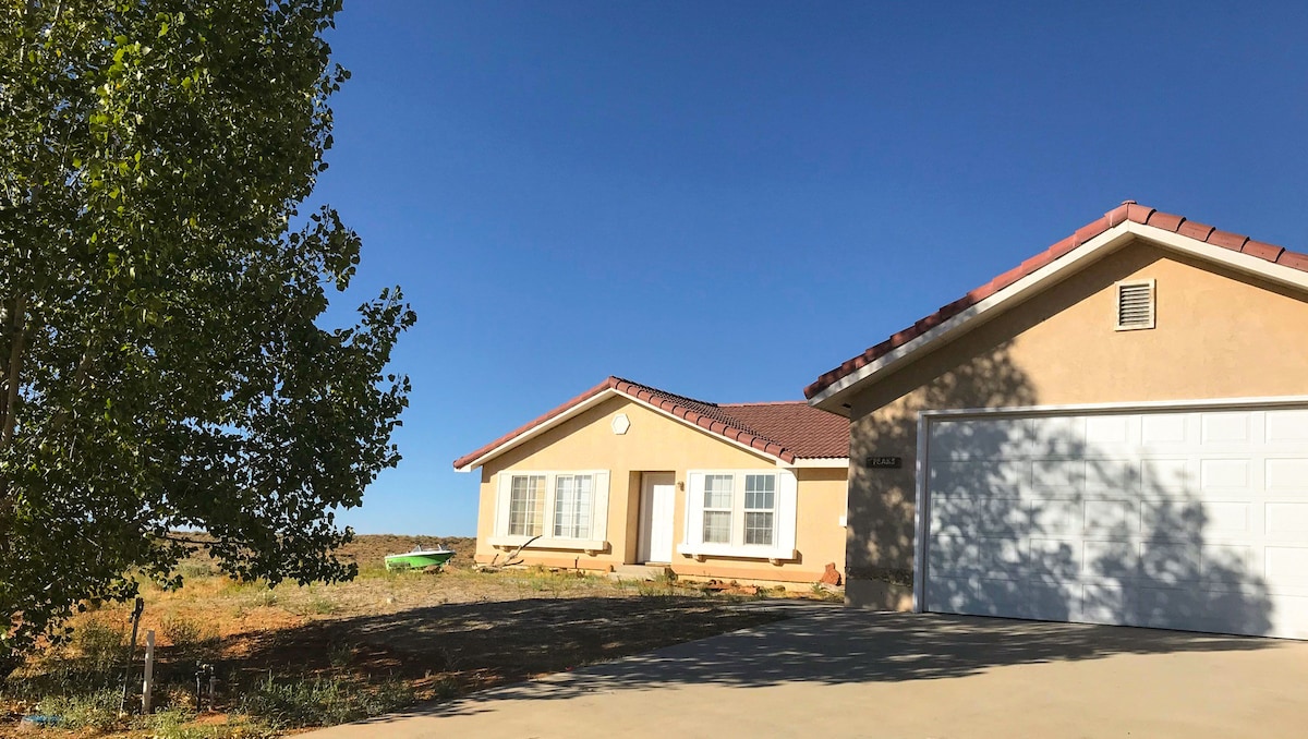 A private home is depicted with a tan exterior and a red-tiled roof, situated against a clear blue sky. The front yard features a level driveway leading to a spacious garage, while patches of greenery are visible along the perimeter.