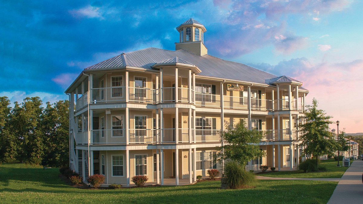The exterior of a three-story building is presented, featuring multiple balconies with railings and a welcoming entrance. Lush green landscaping surrounds the structure, complemented by a vibrant sky with soft clouds during sunset.