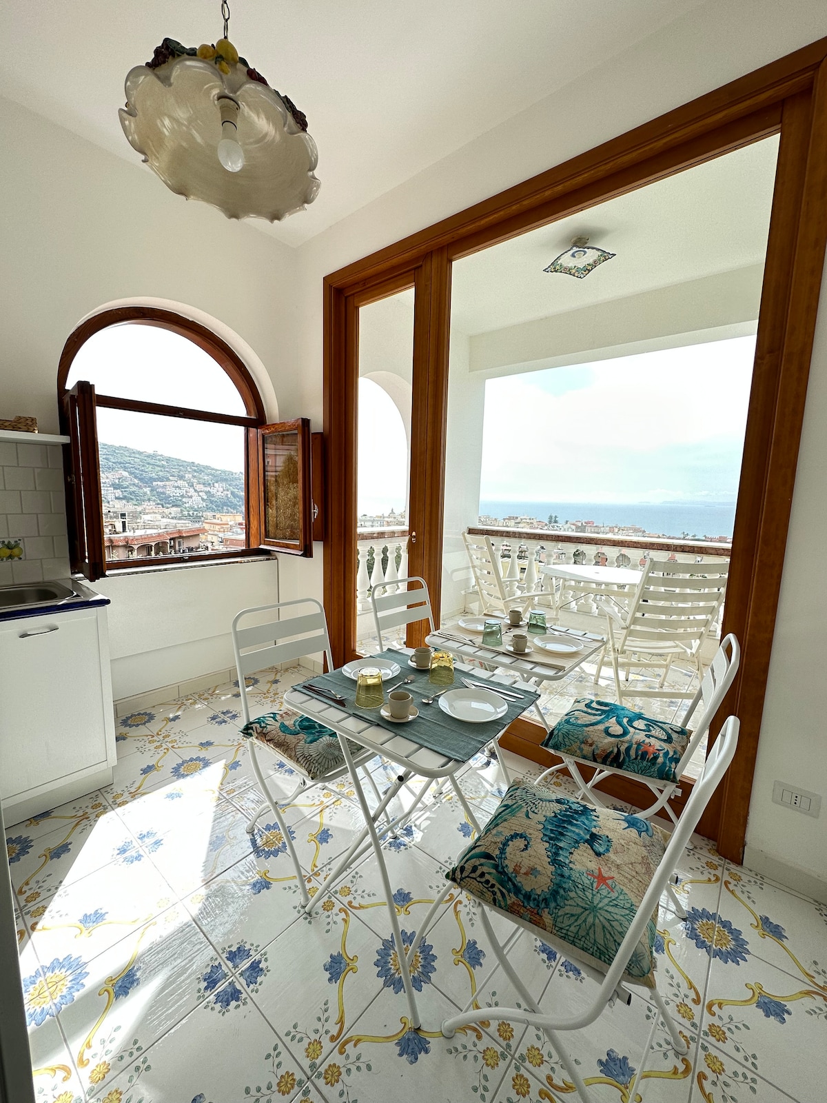 A sunlit dining area features white metal chairs with colorful cushions surrounding a table set for breakfast. Large windows provide panoramic views of the surrounding landscape, with a patterned tiled floor adding a touch of elegance. The atmosphere conveys a bright and airy feel.