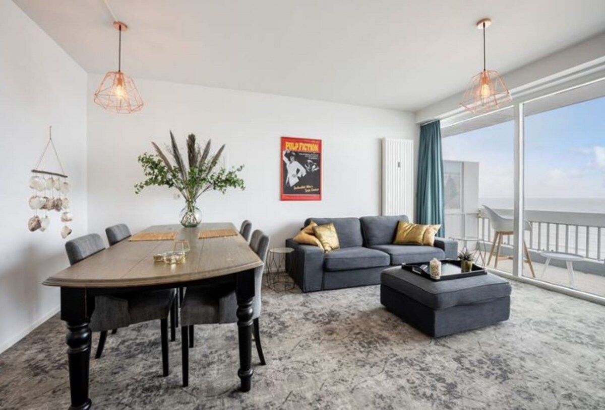 A spacious living area is designed for comfort, featuring a large sectional sofa and a coffee table. A dining table sits nearby with six upholstered chairs. Natural light is provided by large windows, offering views of the sea and direct access to a balcony.
